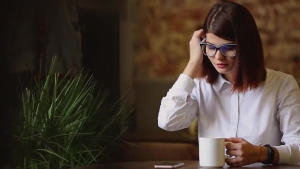 Morena simpática con gafas de primer plano. Bebe café fragante y el uso de un teléfono móvil marca mensajes de texto, ve noticias, hojea fotos, se comunica en una red social . — Vídeo de stock