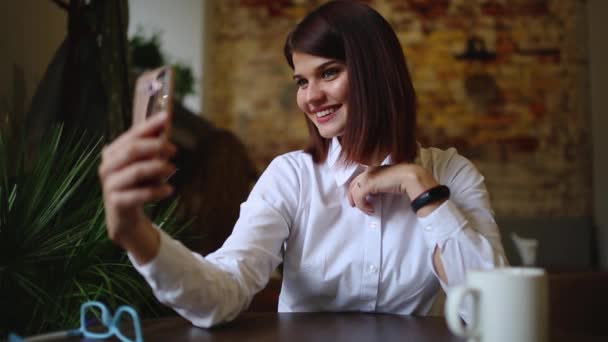 En vacker flicka sitter på ett café och efter druckit en varm kaffe, poserar för en mobiltelefon-kamera för att göra en vacker själv för sociala nätverk. — Stockvideo