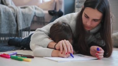 Oğlumla işaretleri ile ailesi tasvir kağıt üzerine çizim yerde yatarken bir sıcak kazaklı güzel genç anne. Çocuk çizmeyi öğrenir. Yakın çekim