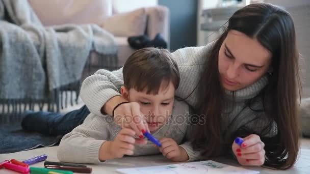 Bela jovem mãe em uma camisola quente deitada no chão com meu filho desenhando com marcadores no papel retratando sua família. A criança aprende a desenhar. Close-up — Vídeo de Stock