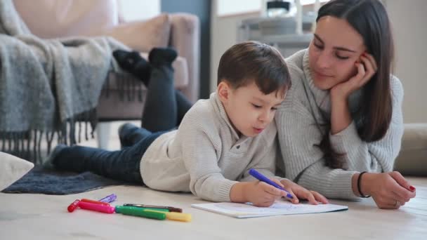 Schöne junge Mutter in einem warmen Pullover auf dem Boden liegend, während mein Sohn mit Filzstiften auf Papier zeichnet, die seine Familie darstellen. das Kind lernt zeichnen. Nahaufnahme — Stockvideo