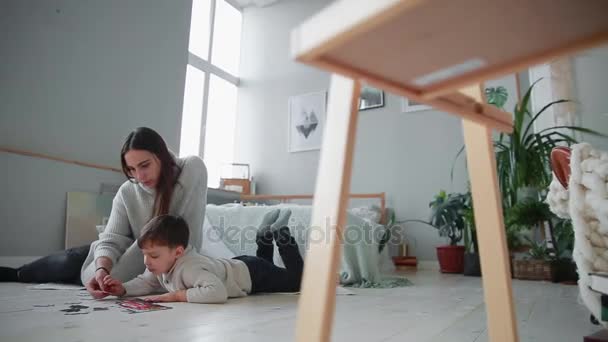 彼の若い息子と一緒にジグソー パズルを収集するために彼の家の白のインテリアで子供を持つ母です。幸せな家庭、教育用ゲーム. — ストック動画