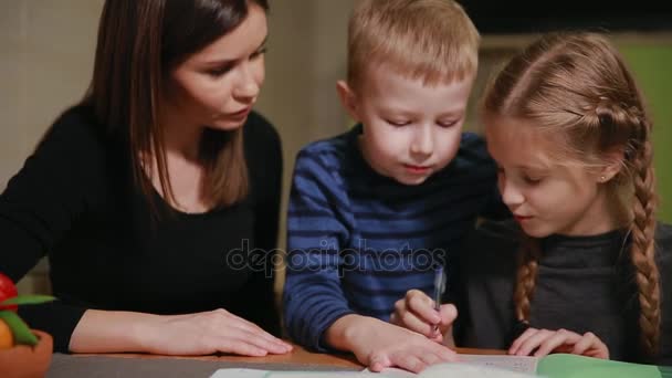 Moeder en dochter doen een huiswerkopdracht school, een kleine jongen horloges het. Broer helpt om huiswerk te doen. — Stockvideo