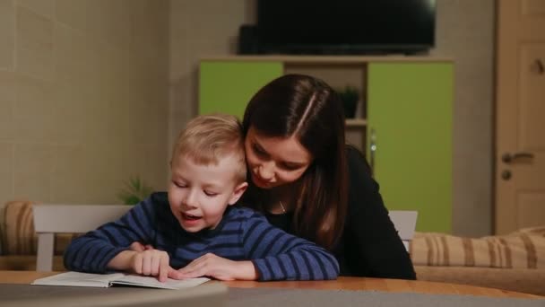 Madre e hijo sentados en la cocina de su casa y aprendiendo a leer el libro . — Vídeo de stock