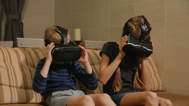 Брат з сестрою вдома сидить на дивані, граючи в гру, використовуючи окуляри віртуальної реальності. Щаслива сім'я — стокове відео