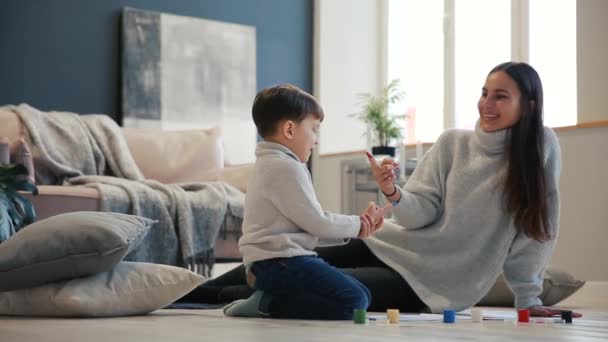 Mother with a child in the white interior of his house was opened the paint can sitting on the floor and dip your fingers in paint and draw on paper with your hands. Happy family. — Stock Video