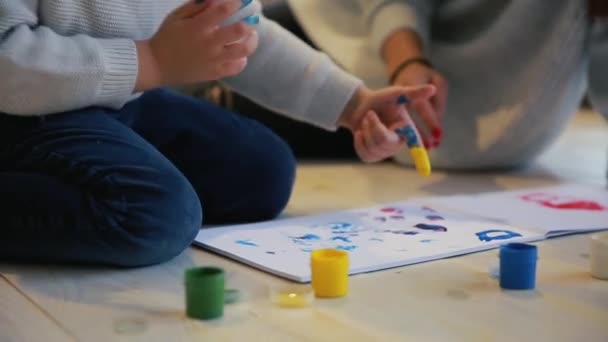 母亲带着一个孩子在他的房子的白色内部被打开了油漆可以坐在地板上, 蘸你的手指在油漆和用手在纸上画画。幸福的家庭。特写 — 图库视频影像