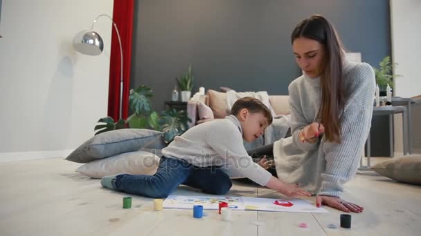 Mother with a child in the white interior of his house was opened the paint can sitting on the floor and dip your fingers in paint and draw on paper with your hands. Happy family. — Stock Video