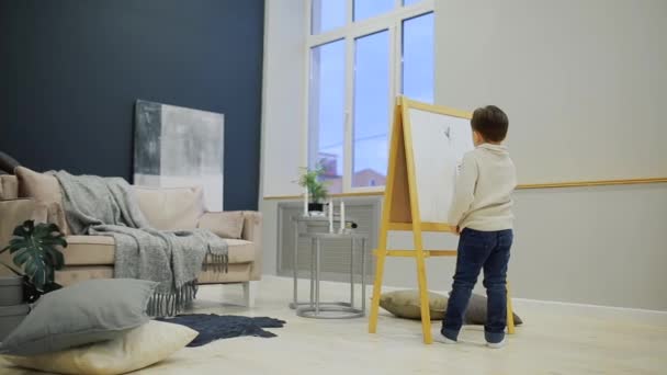 Der kleine Junge im Wohnzimmer des Hauses zeichnet auf dem Brett mit Filzstiften eine Kinderzeichnung. Vorschulbildung. — Stockvideo