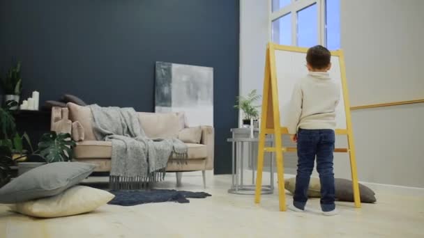 Der kleine Junge im Wohnzimmer des Hauses zeichnet auf dem Brett mit Filzstiften eine Kinderzeichnung. Vorschulbildung. — Stockvideo