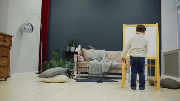 Der kleine Junge im Wohnzimmer des Hauses zeichnet auf dem Brett mit Filzstiften eine Kinderzeichnung. Vorschulbildung. — Stockvideo