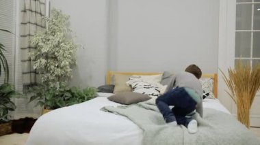 Çocuk 5-7 yıl eski kot ve mutlu bir oyuncak ayı ve yastık ile ebeveyn yatak odasında yatağın üstüne atlama bir gri kazak Avrupa görünüm.