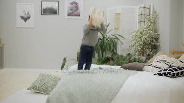 Çocuk 5-7 yıl eski kot ve mutlu bir oyuncak ayı ve yastık ile ebeveyn yatak odasında yatağın üstüne atlama bir gri kazak Avrupa görünüm.