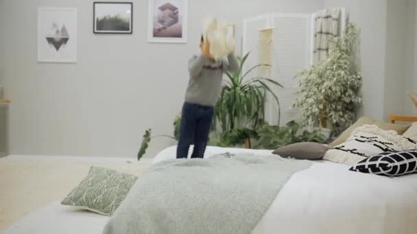 Niño de 5-7 años en vaqueros azules y un suéter gris aspecto europeo saltando felizmente en la cama en el dormitorio de los padres con un oso de juguete y almohada . — Vídeos de Stock