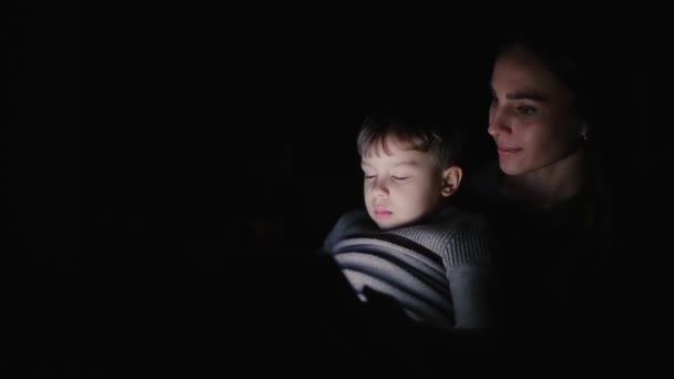 Piękna kobieta matka siedzi w fotelu swojego domu z synem na kolanach oglądać kreskówki na ekranie dotykowym tabletu. — Wideo stockowe