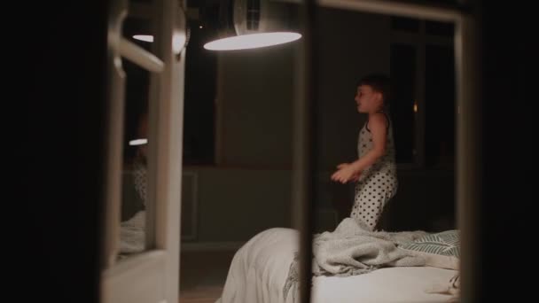 Junge im Schlafanzug springt auf das Bett im Schlafzimmer seines Hauses. Lachen spielt mit einem Spielzeug. — Stockvideo