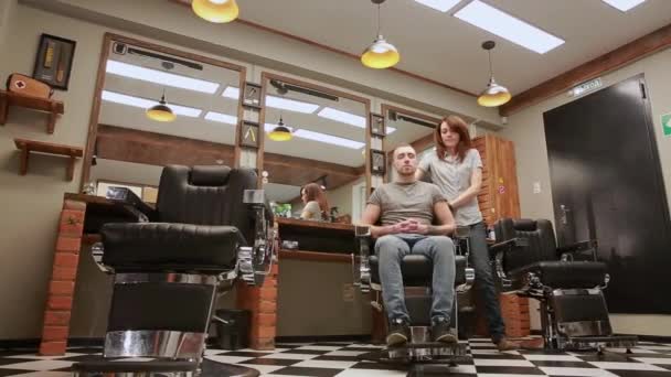 Женщина парикмахер в парикмахерской, чтобы посадить покупателя в кресло и начинает проводить его стрижку. Значение — стоковое видео