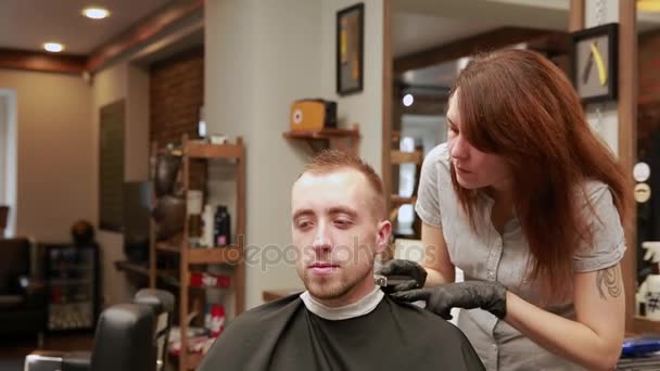 Una mujer barbero en la barbería para poner clientes hombre en una silla y comienza a llevar a cabo su corte de pelo . — Vídeos de Stock