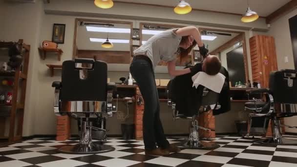 La mujer es una peluquera profesional baja la silla, la cámara se mueve con la ayuda de imágenes de HD Steadicam prístina. Peluquero en la barbería comienza a afeitar al hombre con la cuchilla . — Vídeos de Stock