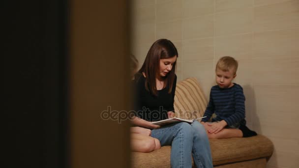 Mama siedzi na kanapie z jego córka i syn, czytanie ich historię, siedząc na kanapie w salonie. — Wideo stockowe