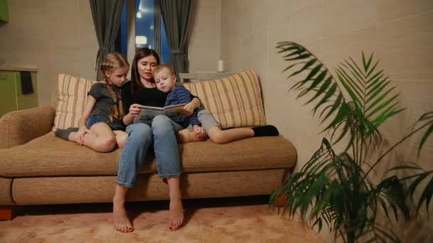 妈妈坐在沙发上与他的女儿和年幼的儿子, 读他们的故事, 而坐在沙发上在他们的客厅. — 图库视频影像