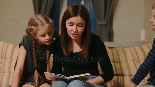 Mom sitting on the couch with his daughter and young son, reading them a story while sitting on the couch in their living room. — Stock Video