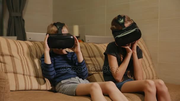 Брат с сестрой дома, сидят на диване и играют в игру, используя очки виртуальной реальности. Счастливая семья — стоковое видео