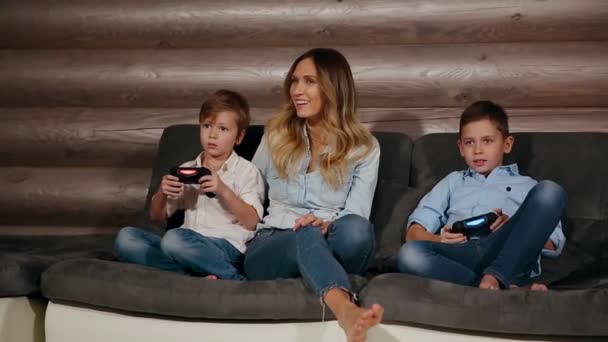 Madre y dos hijos sentados en el sofá de su casa jugando videojuegos con joystick inalámbrico. Gente feliz en la casa . — Vídeos de Stock