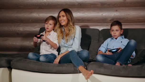 母とワイヤレス ジョイスティックでビデオ ゲームをプレイする彼の家のソファーに座っている 2 人の息子。家の中で幸せな人. — ストック動画