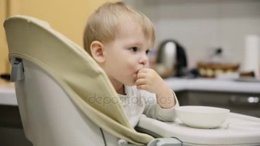 Küçük çocuk mutfakta ve tabakaları gelen çocuk koltuğunda oturan çok lezzetli yiyecek var. Gülümsüyor ve o dönemin oyunları.