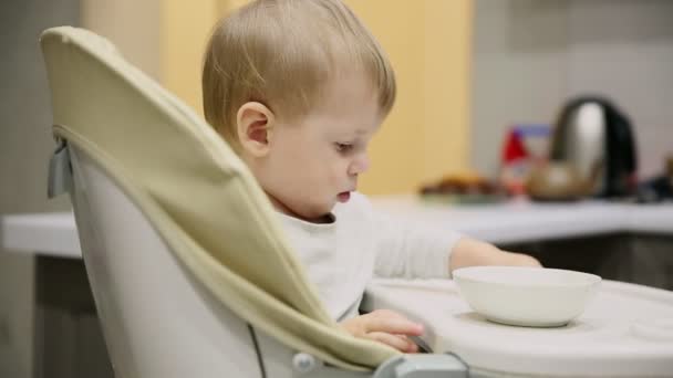 Petit garçon assis dans le siège enfant dans la cuisine et dans les assiettes est une nourriture très savoureuse. Sourires et jeux . — Video