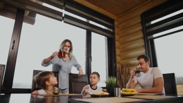 Familia de cuatro desayunando en su cocina con grandes ventanales. La gente sonríe, la madre besa y abraza a los niños. Madre padre y dos hijos . — Vídeos de Stock