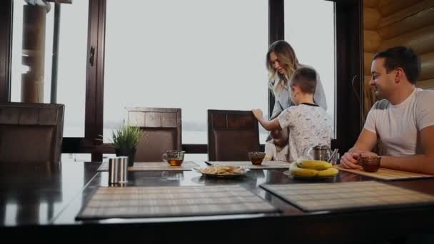 Familia de cuatro desayunando en su cocina con grandes ventanales. La gente sonríe, la madre besa y abraza a los niños. Madre padre y dos hijos . — Vídeos de Stock