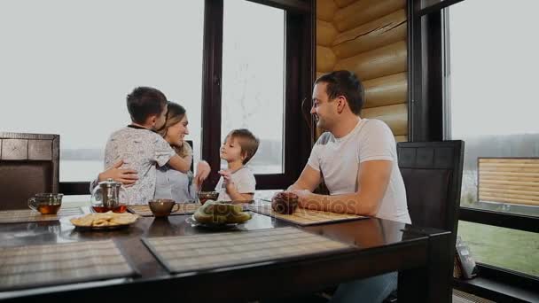 Familj på fyra med frukost i hans kök med stora fönster. Människor ler, mor kyssas och kramas barn. Mor far och två barn. — Stockvideo