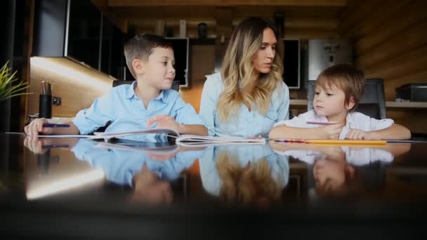 Счастливая мать семьи двоих детей помогает сыновьям делать домашнее задание сидя за большим столом на кухне . — стоковое видео