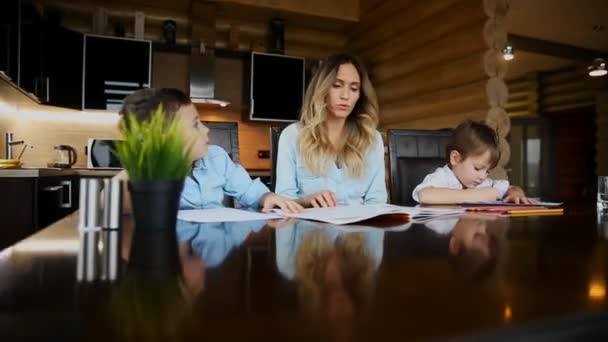 두 아이의 행복 가족 어머니 도움이 부엌 큰 테이블에 앉아 그들의 숙제를 할 아들. — 비디오