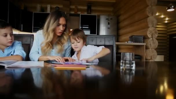 Feliz familia madre de dos hijos ayuda a los hijos a hacer su tarea sentados en la mesa grande en la cocina . — Vídeo de stock