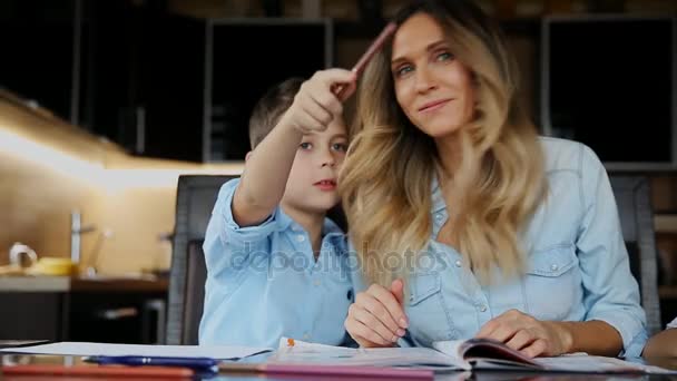 宿題をやって彼の息子を助けて笑顔の母台所のテーブルに座ってを書くためにテキストを指示します。. — ストック動画