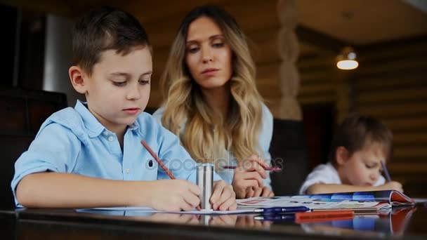 2 人の子供の幸せな家族母親助ける台所の大きなテーブルに座っている彼らの宿題をする息子. — ストック動画