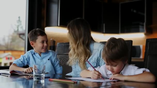 Feliz familia madre de dos hijos ayuda a los hijos a hacer su tarea sentados en la mesa grande en la cocina . — Vídeo de stock