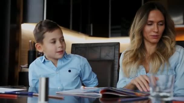 Sonriente madre ayudando a su hijo haciendo la tarea, dicta el texto para ayudarle a escribir sentado en la mesa en la cocina . — Vídeo de stock