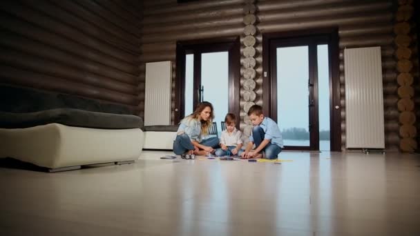 Mãe feliz com dois filhos sentados no chão de sua casa de campo passando tempo juntos coletando crianças designer. Sala de estar com janelas panorâmicas . — Vídeo de Stock