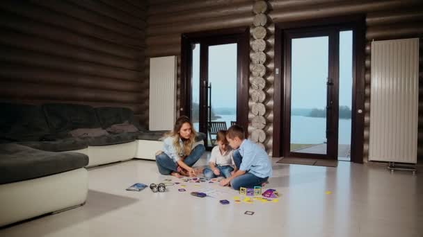 Счастливая мать с двумя детьми, сидящими на полу своего загородного дома, проводит время вместе, собирая детского дизайнера. Гостиная с панорамными окнами . — стоковое видео