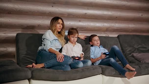 Мать и два сына сидят на диване в его доме и играют в видеоигры с беспроводным джойстиком. Счастливые люди в доме . — стоковое видео