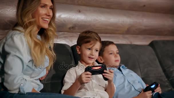 Mother and two sons sitting on the sofa in his house playing video games with wireless joystick. Happy people in the house. — Stock Video