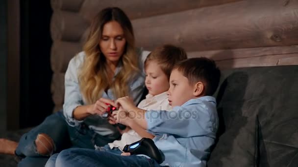 Moeder en twee zonen zittend op de Bank in zijn huis spelen van videospellen met draadloze joystick. Gelukkige mensen in het huis. — Stockvideo