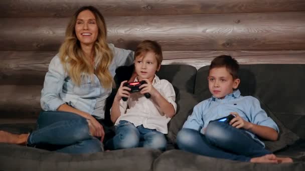 母とワイヤレス ジョイスティックでビデオ ゲームをプレイする彼の家のソファーに座っている 2 人の息子。家の中で幸せな人. — ストック動画