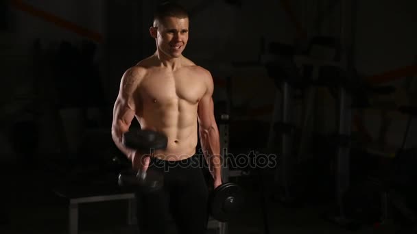 Спортивный юноша без рубашки - модель фитнеса держит гантель в спортзале. Крупный план — стоковое видео