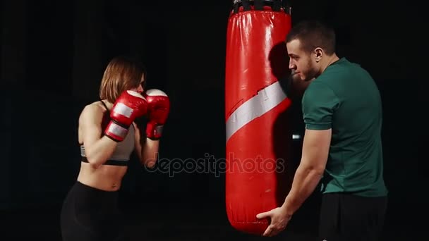 Joven hermosa morena en ropa deportiva está entrenando con su entrenador de boxeo. Sostiene golpes con guantes de boxeo en bolsa sobre fondo negro . — Vídeo de stock