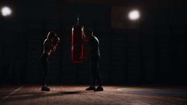 Giovane bella donna bruna in abbigliamento sportivo si allena con il suo allenatore di boxe. Contiene pugni con guanti da boxe in sacchetto su sfondo nero . — Video Stock